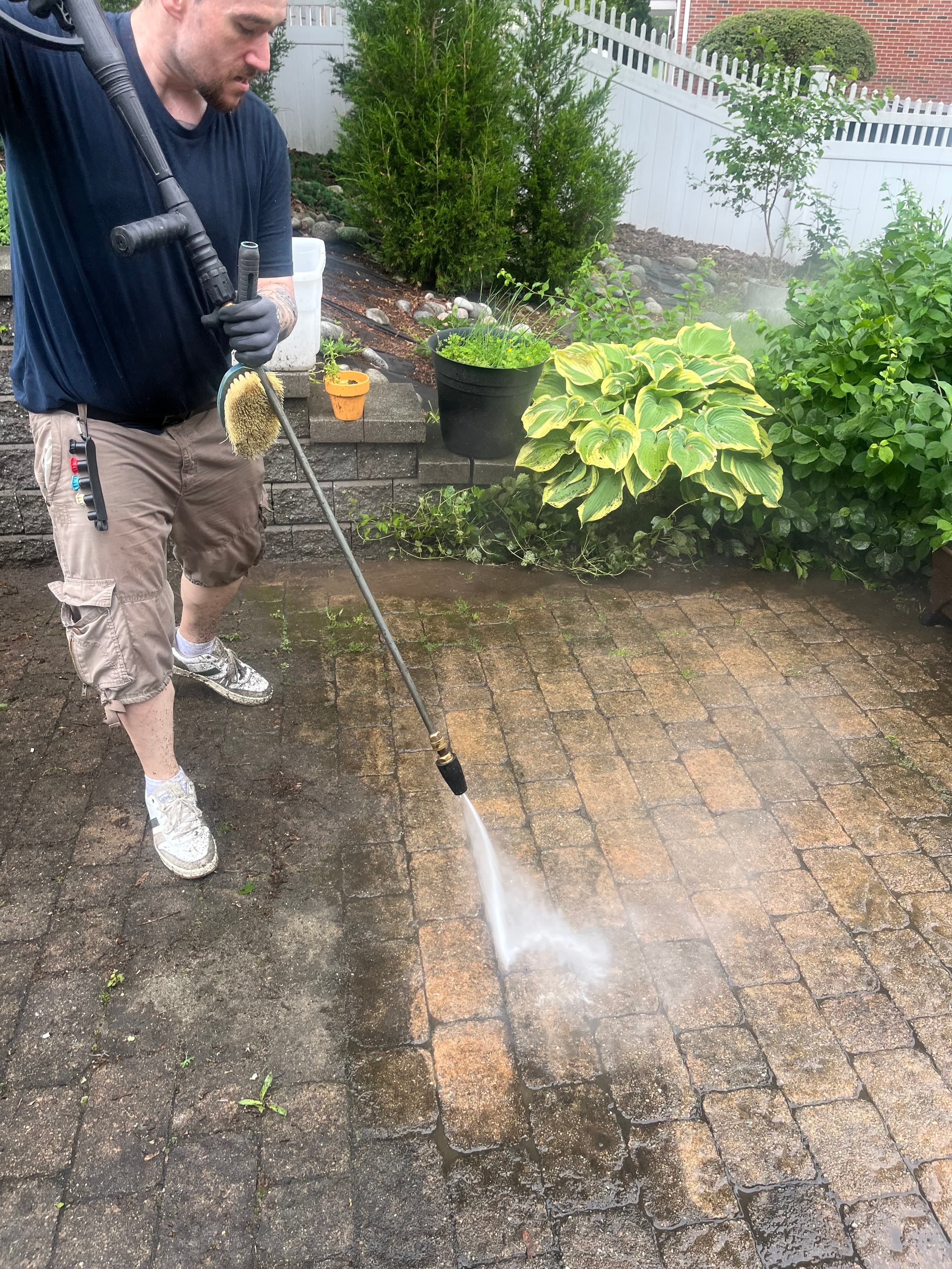 Man pressure washing a stone patio, revealing clean bricks beneath a layer of dark dirt.